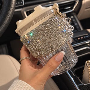 Op de afbeelding: Een doorzichtige plastic beker met een beige deksel en een brede band van fonkelende zilveren strass-steentjes rond het midden. De beker is voorzien van een doorzichtig acryl kettinghandvat bevestigd aan een goudkleurige ring op het deksel.