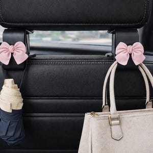 May include: Black car seat headrest hooks with pink bow accents. One hook holds a rolled-up umbrella, the other a light-colored handbag. The hooks are designed to organize items in a vehicle.