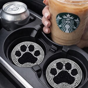 May include: Two black car coasters with a paw print design, embellished with sparkling rhinestones. The coasters sit in a car's cup holder, next to a can and a Starbucks iced coffee.
