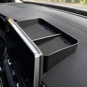 Puede incluir: Una bandeja de almacenamiento negra para salpicadero, de forma rectangular y con un divisor central. La base interior presenta un patrón de rejilla texturizado y antideslizante. La bandeja se sitúa detrás de la pantalla de infoentretenimiento.