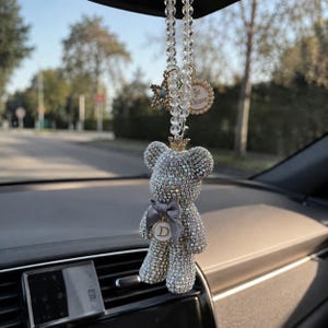Op de afbeelding: Een auto-achteruitkijkspiegelhanger met een berenfiguur bedekt met heldere strass-steentjes, voorzien van een kleine gouden kroon en een grijze strik. Een ronde bedel met de letter D, een sterbedel en een Je t'aime-bedel hangen aan een kralenketting.