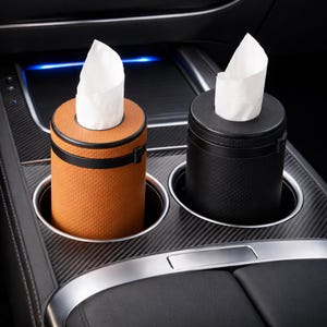 May include: Two car tissue holders, one tan and one black, each holding a stack of white tissues. The holders are cylindrical and fit into car cup holders. The tan holder has a perforated texture.