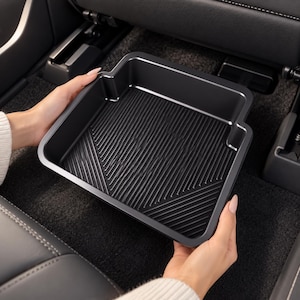 May include: A square black automotive floor tray featuring a raised edge and a textured surface with diagonal grooves. The tray is designed with a specific cutout shape on one side to accommodate the interior contours of a vehicle floor.