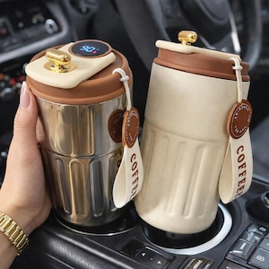 May include: Two insulated coffee tumblers. One is stainless steel with a digital temperature display, the other is cream-colored. Both have brown lids, gold accents, and "COFFEE" straps with leather tags. The tumblers are in a car's console.