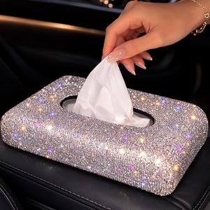 Op de afbeelding: Een rechthoekige tissuebox bedekt met glinsterende strass-steentjes. De box is zilverkleurig met iriserende accenten en een zwarte ovale opening. Witte tissues zijn zichtbaar. De box staat op een zwart oppervlak.