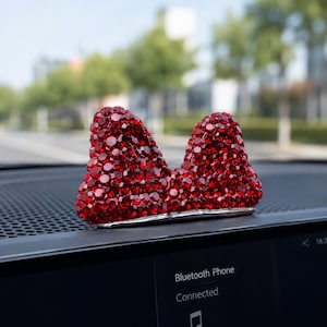 Puede incluir: Un accesorio decorativo para coche con forma de lazo, cubierto totalmente por pequeños cristales rojos facetados. El lazo está situado sobre el salpicadero de un vehículo y cuenta con una base de metal plateado que sostiene la estructura roja.