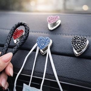 Op de afbeelding: Hartvormige autohaken in rood, roze, blauw en zwart. De zilverkleurige haken zijn versierd met strass-steentjes. Eén haak is bevestigd aan een zwarte gevlochten riem, andere houden witte maskerriemen vast. De haken zijn op een zwart dashboard gemonteerd.