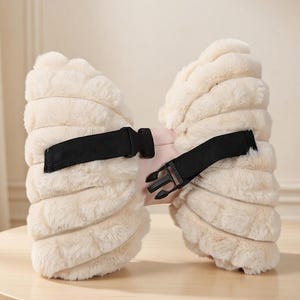 May include: A cream-colored, fluffy bow-shaped accessory with a black strap and buckle. The bow has a soft, textured appearance and a pink center. The black strap is adjustable and secures the accessory.