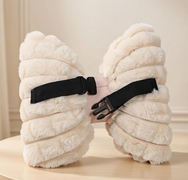 May include: A cream-colored, fluffy bow-shaped accessory with a black strap and buckle. The bow has a soft, textured appearance and a pink center. The black strap is adjustable and secures the accessory.
