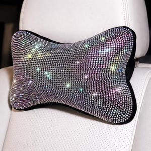 May include: A sparkling car headrest pillow covered in iridescent rhinestones. The pillow has a black border and is shaped like a bone. The pillow is placed on a white car seat.