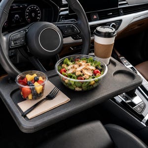 May include: A black plastic car steering wheel tray featuring a recessed cup holder and a flat surface. The tray holds a clear plastic bowl of salad with chicken and tomatoes, a smaller bowl of fruit, a black plastic fork, and a coffee cup.