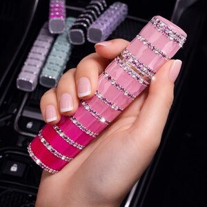 Puede incluir: Un tubo de l&aacute;piz labial rosa y magenta con un dise&ntilde;o degradado, adornado con pedrer&iacute;a. Varios otros tubos de varios colores son visibles en el fondo. El tubo de l&aacute;piz labial se sostiene en una mano con u&ntilde;as cuidadas.