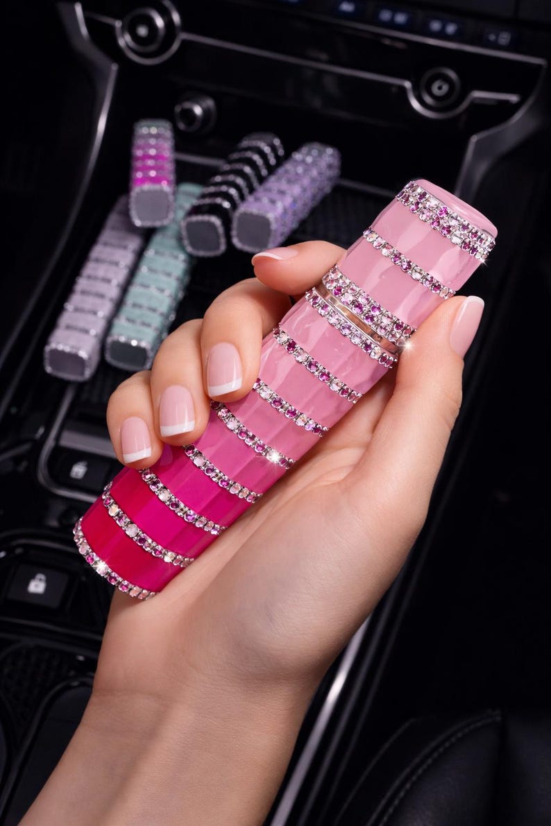 Puede incluir: Un tubo de l&aacute;piz labial rosa y magenta con un dise&ntilde;o degradado, adornado con pedrer&iacute;a. Varios otros tubos de varios colores son visibles en el fondo. El tubo de l&aacute;piz labial se sostiene en una mano con u&ntilde;as cuidadas.