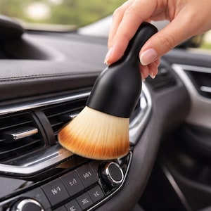 May include: A black car detailing brush with a rounded handle and soft, light-colored bristles with an orange-brown gradient. The brush is being used to clean a car's dashboard, specifically the air vents and control panel.