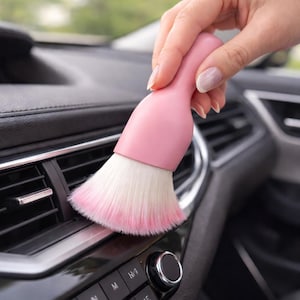 May include: A pink car detailing brush with white and pink bristles is held over a car's dashboard vent. The brush has a rounded handle and is designed for cleaning vehicle interiors.