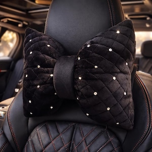 May include: Black bow-shaped car headrest pillow. The pillow is made of a soft, quilted material and is adorned with small, sparkling rhinestones. The headrest is designed to attach to a car seat and provides support for the head and neck.