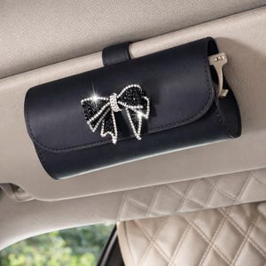 May include: Black car visor sunglasses holder with a decorative bow. The bow is embellished with black and clear rhinestones. The holder is attached to the car visor with a black strap.