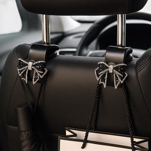 Puede incluir: Ganchos negros para reposacabezas de asiento de coche con detalles de lazo decorativos. Cada gancho presenta un lazo negro adornado con cristales brillantes. Un paraguas negro y una bolsa de la compra con asas negras cuelgan de los ganchos.