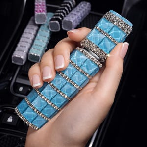 Puede incluir: Un ambientador rectangular azul claro para coche con un patr&oacute;n de diamantes y detalles de pedrer&iacute;a. Varios ambientadores de otros colores son visibles en el fondo.