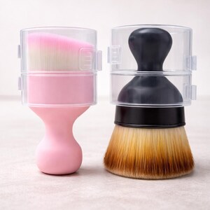 May include: Two makeup brushes with clear protective covers. One brush has a pink handle and a pink and white brush head. The other brush has a black handle and a brown and yellow brush head. Both brushes are on a light-colored surface.