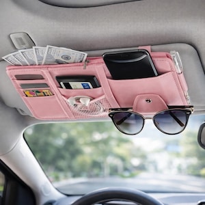 Könnte beinhalten: Ein rosa Auto-Sonnenblenden-Organizer mit mehreren Taschen, die Bargeld, ein Smartphone, Karten und andere kleine Gegenstände enthalten. Sonnenbrillen sind unten befestigt. Der Organizer ist für die Befestigung an der Sonnenblende eines Autos konzipiert.