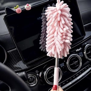 Peut inclure: Un plumeau de voiture en microfibre rose avec un manche rose assorti est tenu devant le tableau de bord d'une voiture. Le plumeau est duveteux et conçu pour nettoyer les surfaces. Des accessoires décoratifs sont fixés à l'écran de la voiture.