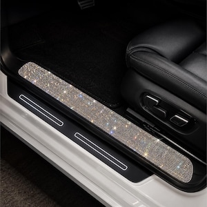 May include: A car door sill protector featuring a long, rectangular strip covered in small, reflective silver rhinestones. The sparkling adhesive strip is applied to the black plastic door entry area of a vehicle, positioned below a black leather seat.