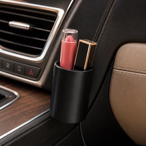 Könnte beinhalten: Ein mattschwarzer, zylindrischer Aufbewahrungsbecher an einer Fahrzeuginnenverkleidung, der einen rosa Lippenstift und ein rechteckiges, schwarz-goldenes Kosmetikgehäuse enthält. Der Becher befindet sich in der Nähe der Lüftung und Holzverkleidung.