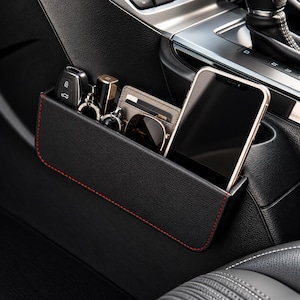 Könnte beinhalten: Ein schwarzer Auto-Sitzlücken-Organizer aus Kunstleder mit roten Kontrastnähten. Die rechteckige Aufbewahrungstasche enthält ein Smartphone, einen Autoschlüssel, einen Lippenstift, eine Sonnenbrille und ein schmales Kartenetui.