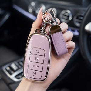 Könnte beinhalten: Ein rosafarbenes Autoschlüsselgehäuse mit einem silberfarbenen Metallrahmen und Knöpfen. Das Schlüsselgehäuse ist an einem silbernen Schlüsselring mit einem lila Lederriemen befestigt. Das Schlüsselgehäuse hat eine glatte, glänzende Oberfläche.