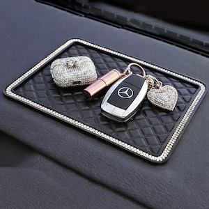 May include: A black, diamond-patterned car dashboard tray with a rhinestone border. Items on the tray include a bedazzled AirPod case, a lipstick, a car key, and a rhinestone heart keychain.