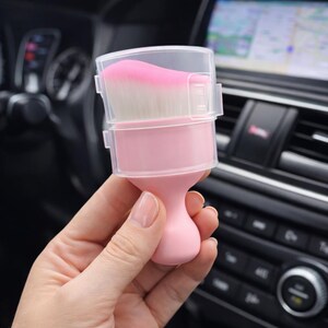 May include: A pink makeup brush with a clear plastic cover. The brush has a rounded handle and a head of pink and white bristles. The brush is held in front of a car's dashboard.