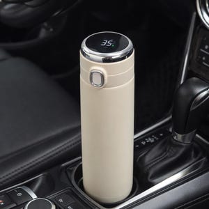 Puede incluir: Una taza de cerámica blanca con un asa negra y el texto "But First, Coffee" en negrita negra. La taza está sobre una superficie blanca. La taza mide 10 cm de alto.