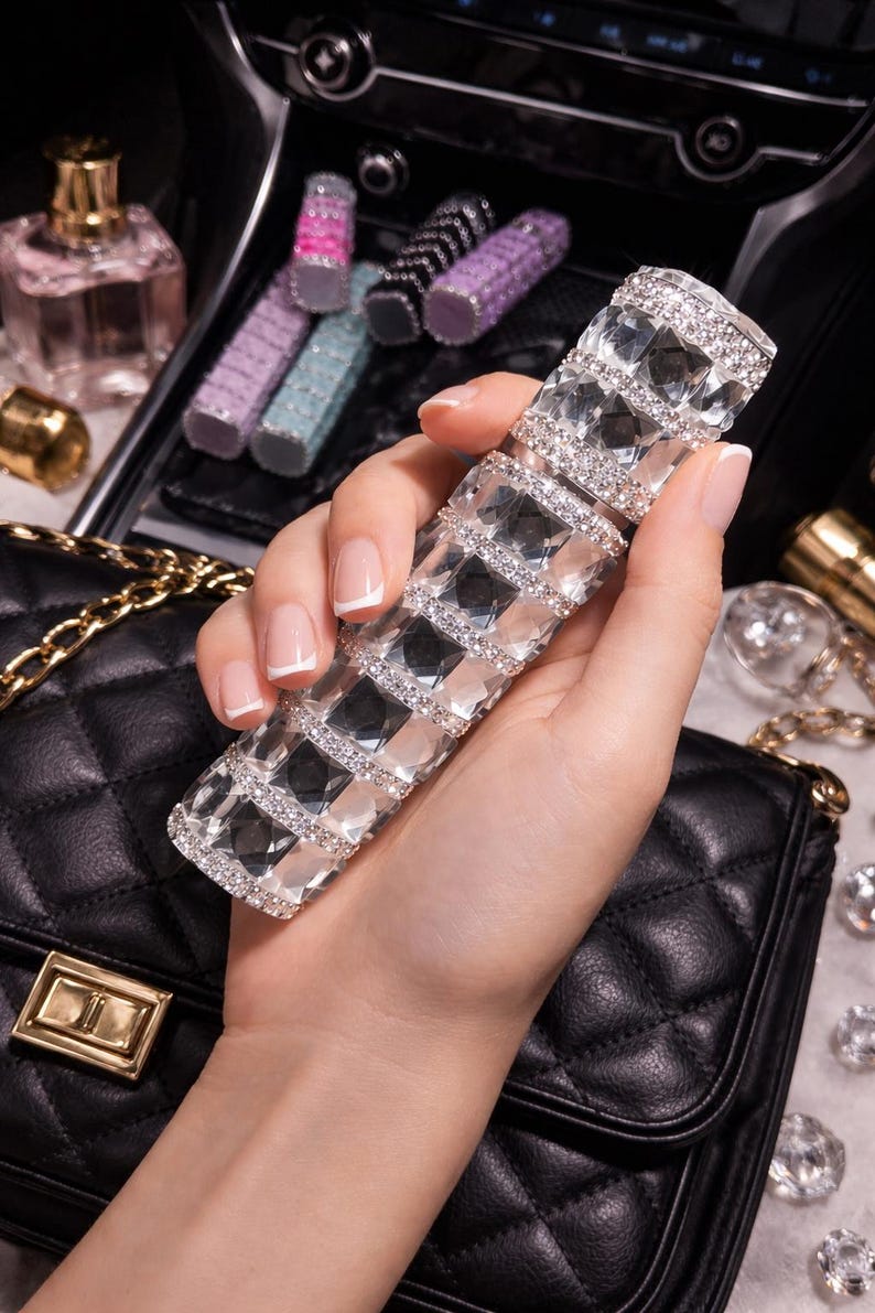 Puede incluir: Un atomizador de perfume rectangular, adornado con cristales, sostenido en una mano. El atomizador es plateado y transparente, con un patr&oacute;n de diamantes. Otros frascos de perfume y barras de labios son visibles en el fondo, junto con un bolso negro acolchado.