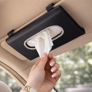 May include: Black faux leather car tissue holder with a rhinestone-lined oval opening. The tissue holder is attached to the car's visor with two black straps. White tissues are visible inside the holder.