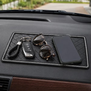 Peut inclure: Un tapis de tableau de bord noir rectangulaire avec un motif en losanges matelassé et une bordure de petits strass argentés. Sur le tapis reposent une clé de voiture noire avec une lanière en cuir tressé, des lunettes de soleil et un smartphone.