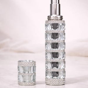 Puede incluir: Un elegante frasco de perfume con un tap&oacute;n a juego. El frasco y el tap&oacute;n est&aacute;n adornados con cristales transparentes y facetados y una parte superior de metal plateado. El dise&ntilde;o es sim&eacute;trico y lujoso, lo que sugiere un producto de fragancia de alta gama.