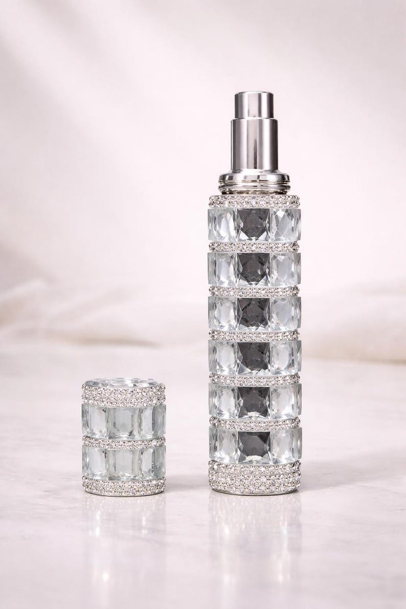 Puede incluir: Un elegante frasco de perfume con un tap&oacute;n a juego. El frasco y el tap&oacute;n est&aacute;n adornados con cristales transparentes y facetados y una parte superior de metal plateado. El dise&ntilde;o es sim&eacute;trico y lujoso, lo que sugiere un producto de fragancia de alta gama.