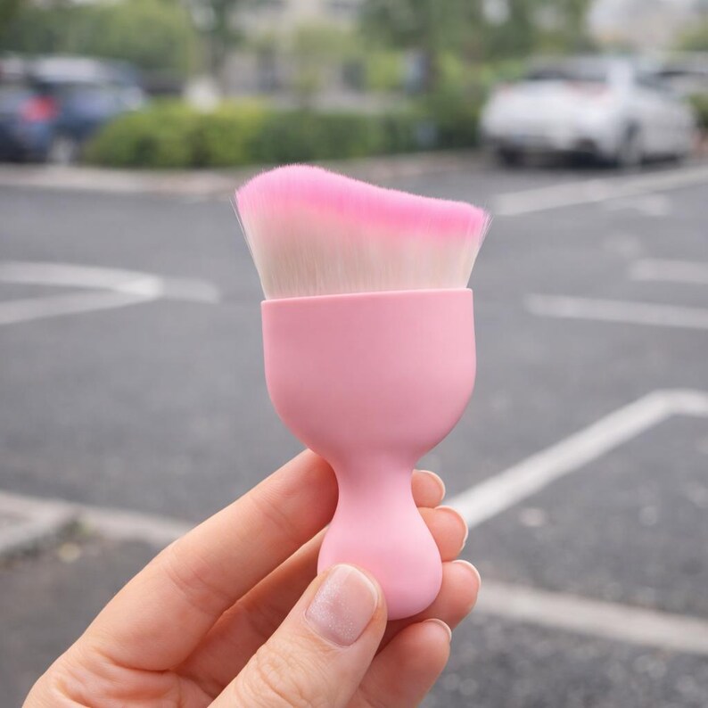 May include: A pink makeup brush with a rounded handle and a flat, angled brush head. The brush head features a gradient of pink to white bristles. The brush is held in a hand against a blurred outdoor background.