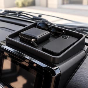 Könnte beinhalten: Eine schwarze, rechteckige Ablageschale für das Auto-Armaturenbrett mit strukturierter Unterlage. Die Schale enthält eine schwarze Geldbörse mit Reißverschluss, eine schwarze Sonnenbrille und ein schwarzes Smartphone auf dem Armaturenbrett.