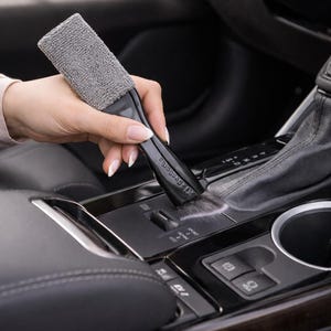 May include: A black car detailing brush with a gray microfiber cleaning head. The brush is being used to clean a car's center console, which is black with silver accents. The interior of the car is black.