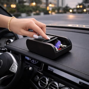 Puede incluir: Una caja de almacenamiento negra para el salpicadero del coche con tapa corredera. La caja está abierta, revelando llaves y otros objetos pequeños. Una mano con una pulsera dorada está abriendo la tapa. El interior del coche es visible.