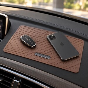 Könnte beinhalten: Eine rechteckige braune Armaturenbrettmatte mit einem sich wiederholenden Fischgrätenmuster und einem kleinen Akzentstück aus Kohlefaser. Ein schwarzer Mercedes-Benz Autoschlüssel und ein dunkelgraues Smartphone liegen auf der Matte auf dem Armaturenbrett.