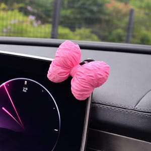 Può includere: Un accessorio per auto a forma di farfalla rosa acceso e peluche. La farfalla è attaccata a una superficie nera, forse un cruscotto dell'auto. Lo sfondo mostra una vista sfocata di vegetazione e un interno dell'auto.