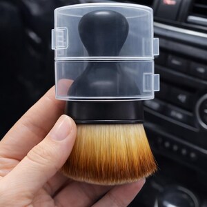 May include: A black makeup brush with a clear plastic cover. The brush has a black handle and a rounded brush head with a gradient of tan and brown bristles. The brush is designed for applying makeup.