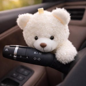 May include: A soft, white teddy bear with a small yellow detail on its head sits on a black car control lever. The bear has a brown nose and dark eyes. The background shows the car's interior.
