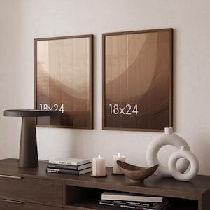 May include: Two 18x24 inch brown wooden picture frames hanging on a wall above a dark wooden sideboard. The sideboard holds a dark brown lamp, two white pillar candles, a wooden bowl, and two white circular vases.