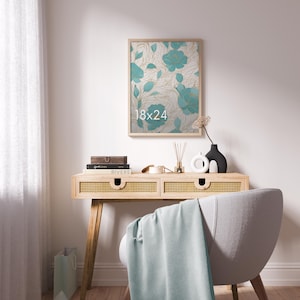 May include: A rectangular wall art print in a light wood frame featuring a teal floral pattern with gold line accents and the text 18x24. The artwork is displayed above a light wood console table with cane drawer fronts and a light blue throw.