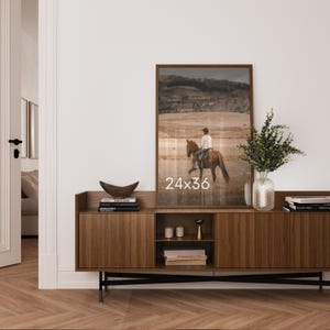 May include: A framed vertical wall art print measuring 24x36 inches, depicting a person riding a brown horse in a vast, dry landscape. The artwork is displayed on a dark wood sideboard with vertical slats, alongside a glass vase with greenery.