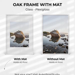 May include: Two white framed art prints, one with a mat and one without, showcasing a landscape scene with a large rock formation in a lake. The text "OAK FRAME WITH MAT" and "Glass • Plexiglass" are at the top. The text "With Mat Holds A2 Print" and "Without Mat Holds A1 Print" are below the frames.